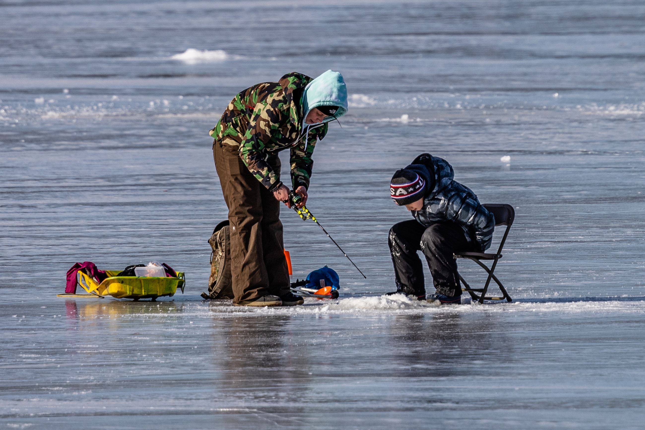 Two young fisherman peer into a hole in a frozen lake, one stands with a fishing rod, the other sits